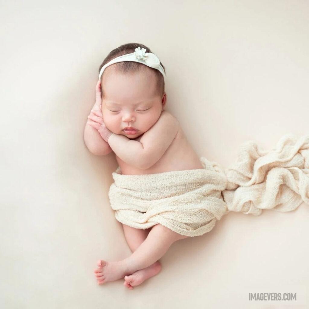 A newborn baby is sleeping peacefully with her head on one hand wrapped in a cream colored net cloth and a hairband around her forehead.jpg 10 11zon