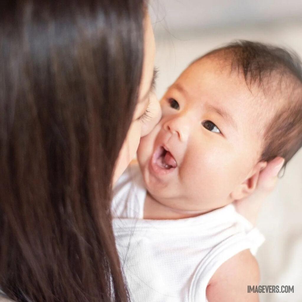 A newborn baby wearing a white cloth in his mothers lap is looking at his mother tearing his mouth and his mother is kissing his left cheek.jpg 26 11zon