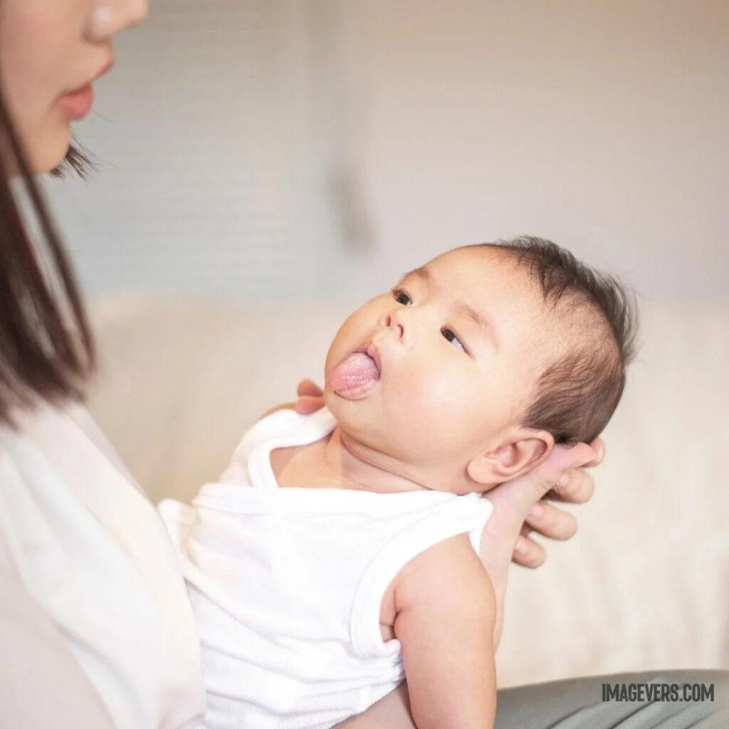 A newborn baby wearing a white cloth on his mothers lap is looking at his mother sticking out her tongue