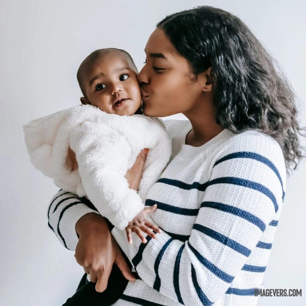 A newborn baby wearing a white cloth on his mothers lap is looking at someone on the right and his mother is lovingly kissing his cheeks.jpg 28 11zon